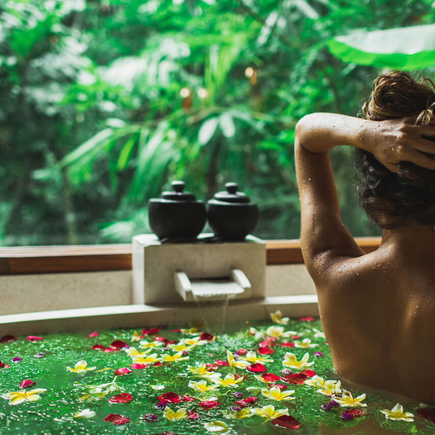 Beautiful young woman enjoying in spa, view from behind. Luxury stone bath tub with jungle view in window. Tropical flowers in water. Beauty treatment concept.